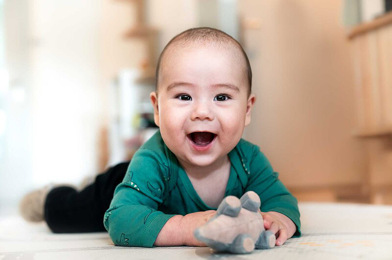 Ein fröhlich lachendes Baby liegt auf dem Boden und hält ein Spielzeug, aufgenommen während eines entspannten Katzenshootings zu Hause in Luzern.