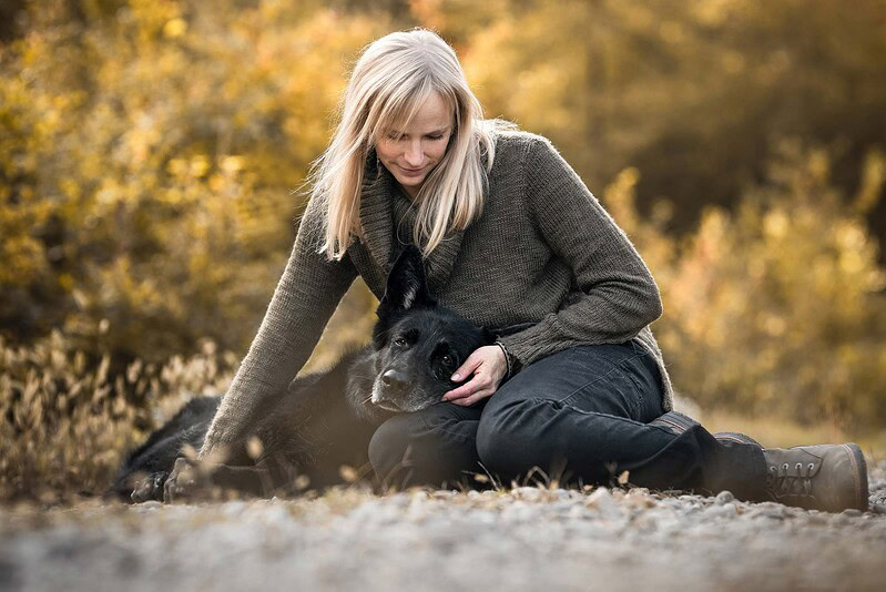 Eine Frau sitzt auf dem Waldboden und hält sanft den Kopf ihres alten schwarzen Schäferhundes mit grauem Gesicht, aufgenommen bei einem Regenbogen-Fotoshooting im Herbst nahe Bern.