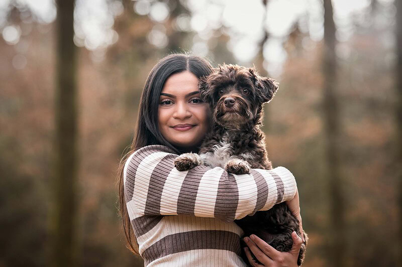 Eine junge Frau mit einem gestreiften Pullover hält einen braunen Bolonka Zwetna im Arm, aufgenommen während eines Herbst-Fotoshootings im Wald bei Deitingen (Solothurn).